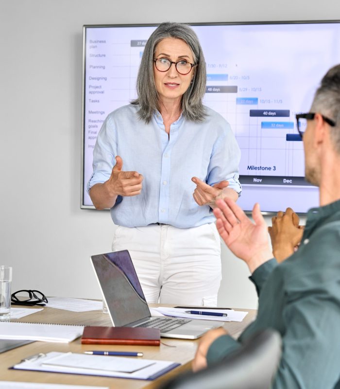 Senior mature older female executive ceo presenter in glasses discussing presentation of corporation financial global goals on big screen with executives at boardroom modern office. Vertical.